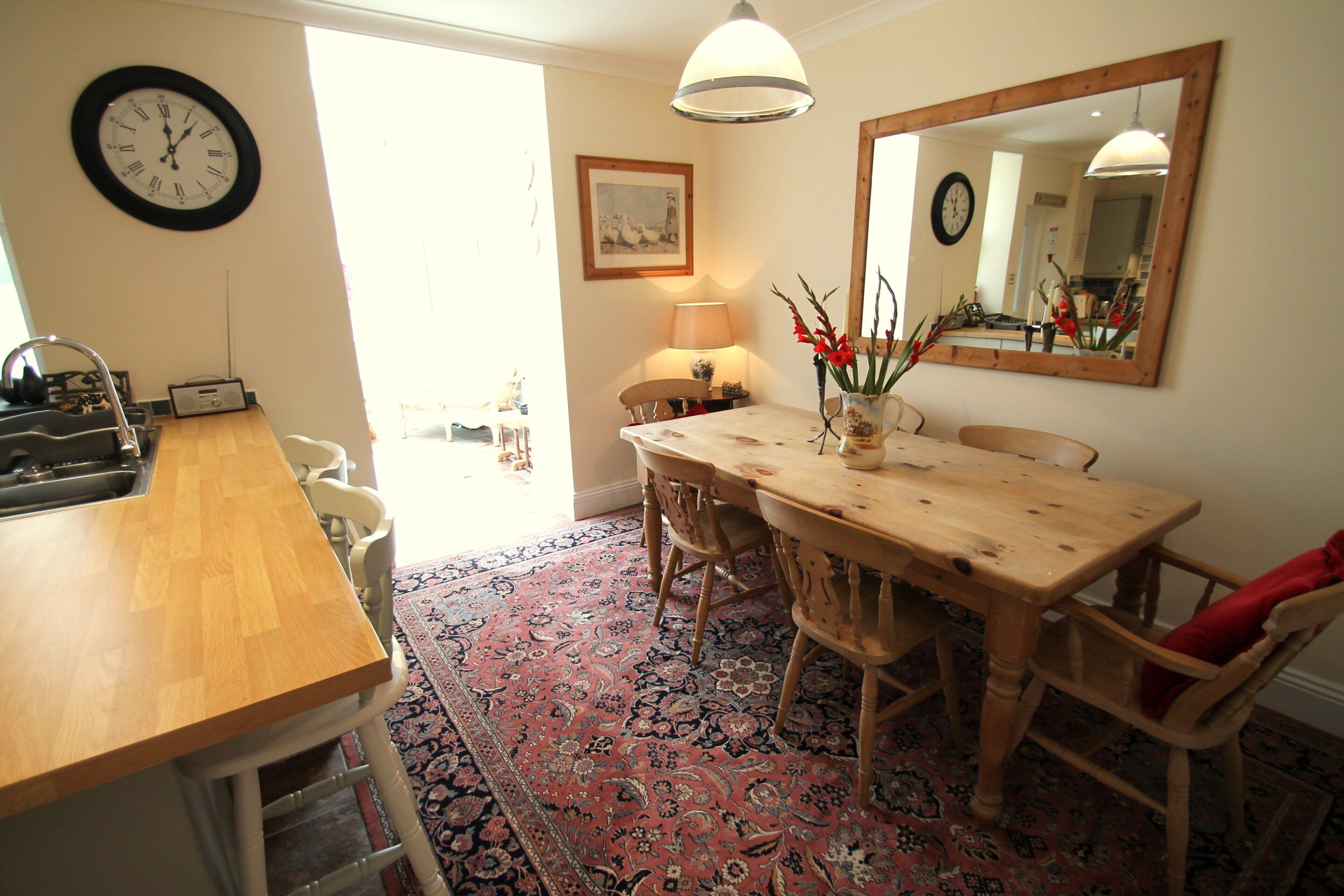 A dining area with a wooden table, chairs, a rug, a clock, and a mirror.