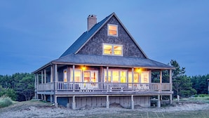A large, two-story wooden house with a balcony, surrounded by natural landscape.
