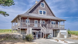 A large, two-story wooden house with a balcony on the second floor, situated on a sandy beach with a clear sky.
