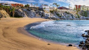 A beach with clear water, rocky shoreline, and buildings in the background.