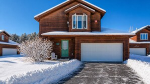 Une maison en bois à deux étages, avec une porte verte, un garage et une allée recouverte de neige.