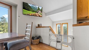 Une pièce comprenant une table à manger en bois, une chaise rembourrée, un téléviseur fixé au mur, une échelle en bois et un panier contenant des produits de nettoyage.