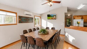 Une salle à manger avec une table en bois, des chaises, un ventilateur de plafond et un coin cuisine équipé de placards et d’un réfrigérateur.