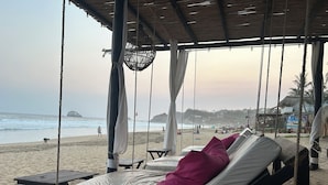 Un salón junto a la playa con techo de paja, asientos acolchados y vista al océano.