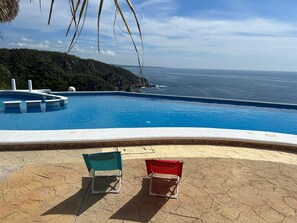 A swimming pool with a view of the ocean, two beach chairs, and a stone patio.