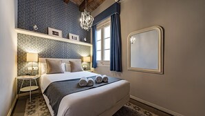 A neatly arranged bedroom with a large bed, bedside lamps, a chandelier, and a view of the outdoors.