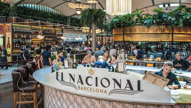 A bustling bar area with a counter labeled "EL NACIONAL BARCELONA," surrounded by patrons and a variety of bottles.