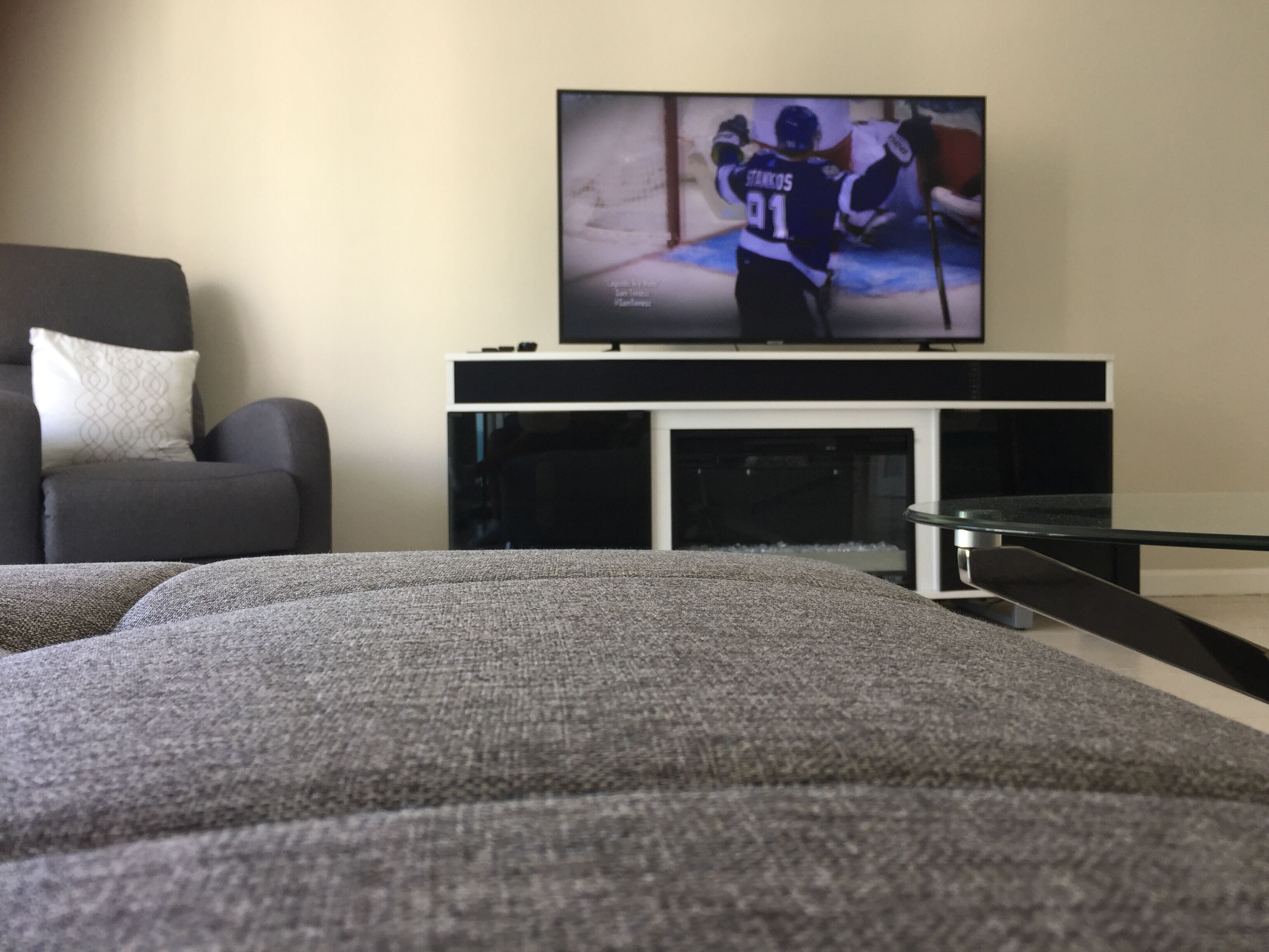 A living room with a flat-screen TV on a black stand, a grey sofa, and a white cushion.