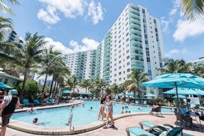 A pool area with lounge chairs and umbrellas, surrounded by palm trees and a tall hotel building.