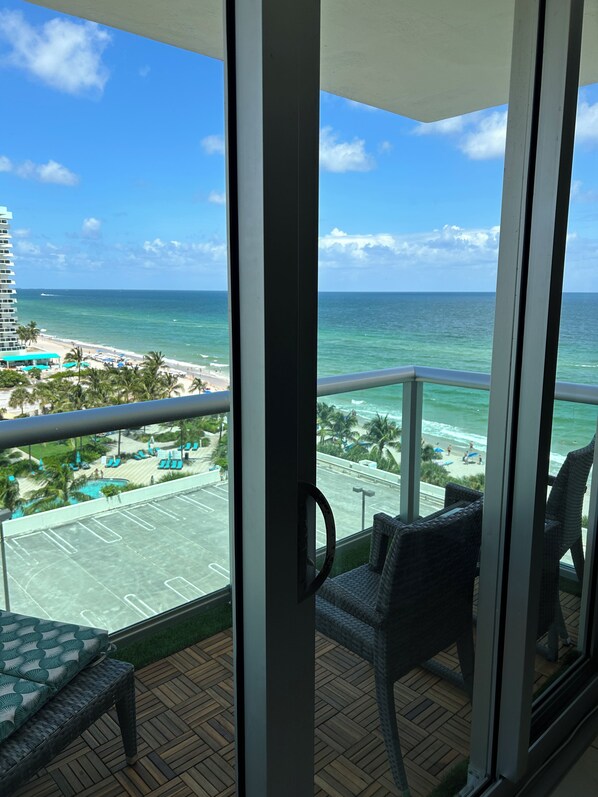 A balcony with a view of the beach and ocean, featuring a pool and lounge chairs.
