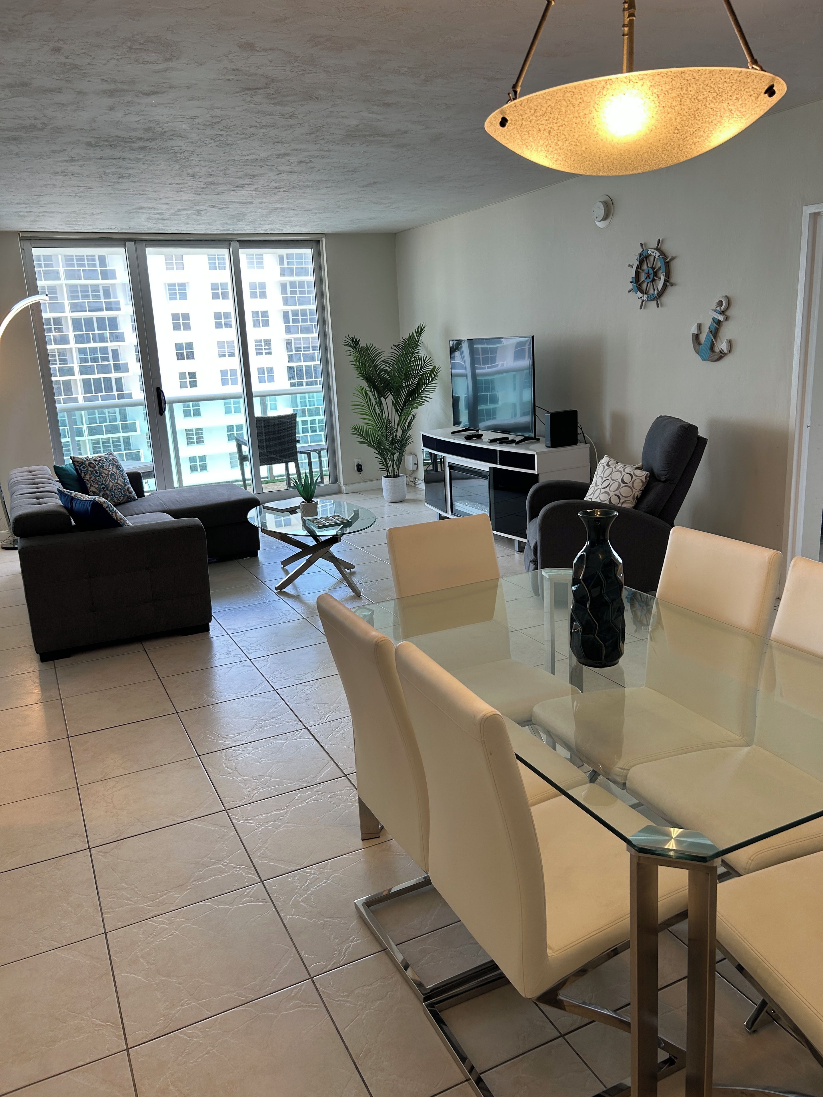 A modern living room with a glass dining table, comfortable seating, a flat-screen TV, and large windows offering a city view.