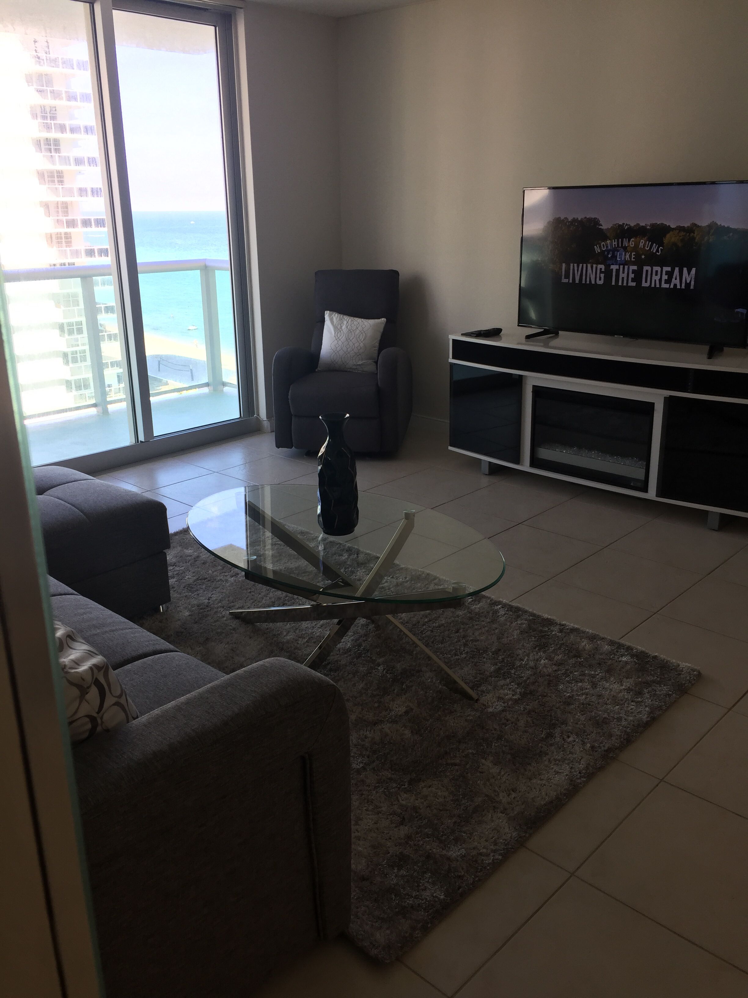A modern living room with a glass coffee table, grey sofa, and a flat-screen TV on a stand.