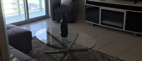 A modern living room with a glass coffee table, grey sofa, and a flat-screen TV on a stand.