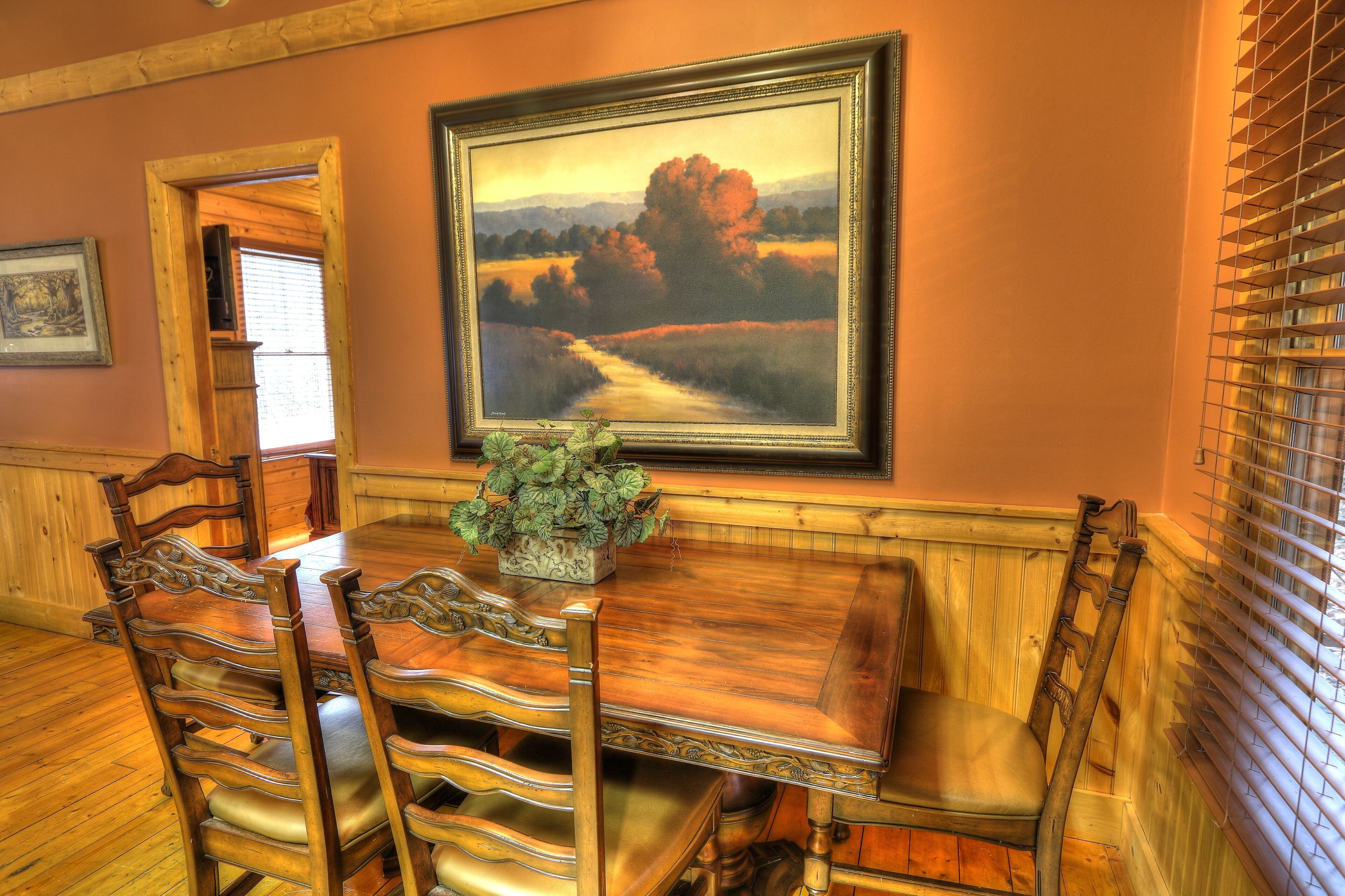 A dining area with a wooden table and chairs, a framed landscape painting, and a potted plant.