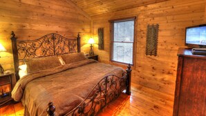 A wooden cabin bedroom with a bed, nightstands, a television, and a window with blinds. - Family Tyme #78 - Charming & Clean Log Cabin! (Sevierville)