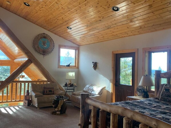 A cozy cabin interior with a sloped ceiling, wooden walls, a sofa, a chair, a small table, and a bed.