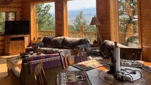 A log cabin interior with a large window, a mounted deer head, a cozy seating area, and a table with a quilt.