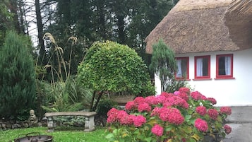 Escape to a serene garden oasis with a charming thatched-roof cottage surrounded by lush greenery and vibrant hydrangea bushes.