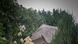 Eine rustikale Holzhütte mit Strohdach, umgeben von einem Kiesweg und Grünflächen.