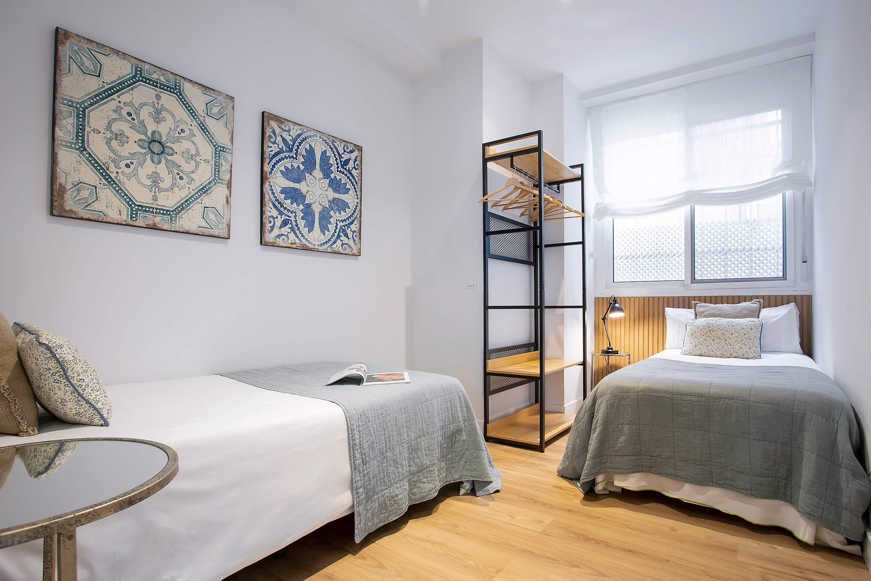 Een slaapkamer met twee bedden, een houten vloer, een raam met witte gordijnen en een metalen schapunit.