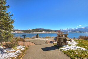 Un sentier pavé mène à un banc et à une structure en pierre, le tout situé au bord d’un lac, avec des montagnes en arrière-plan.