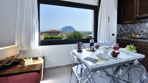 A modern kitchen with a large window, a dining area with a table and chairs, and a view of a mountain and buildings. - Holiday apartments L'Ea di Lavru 
SEA VIEW 3-rooms apartment-TOP INTERIOR DESIGN (Porto San Paolo)