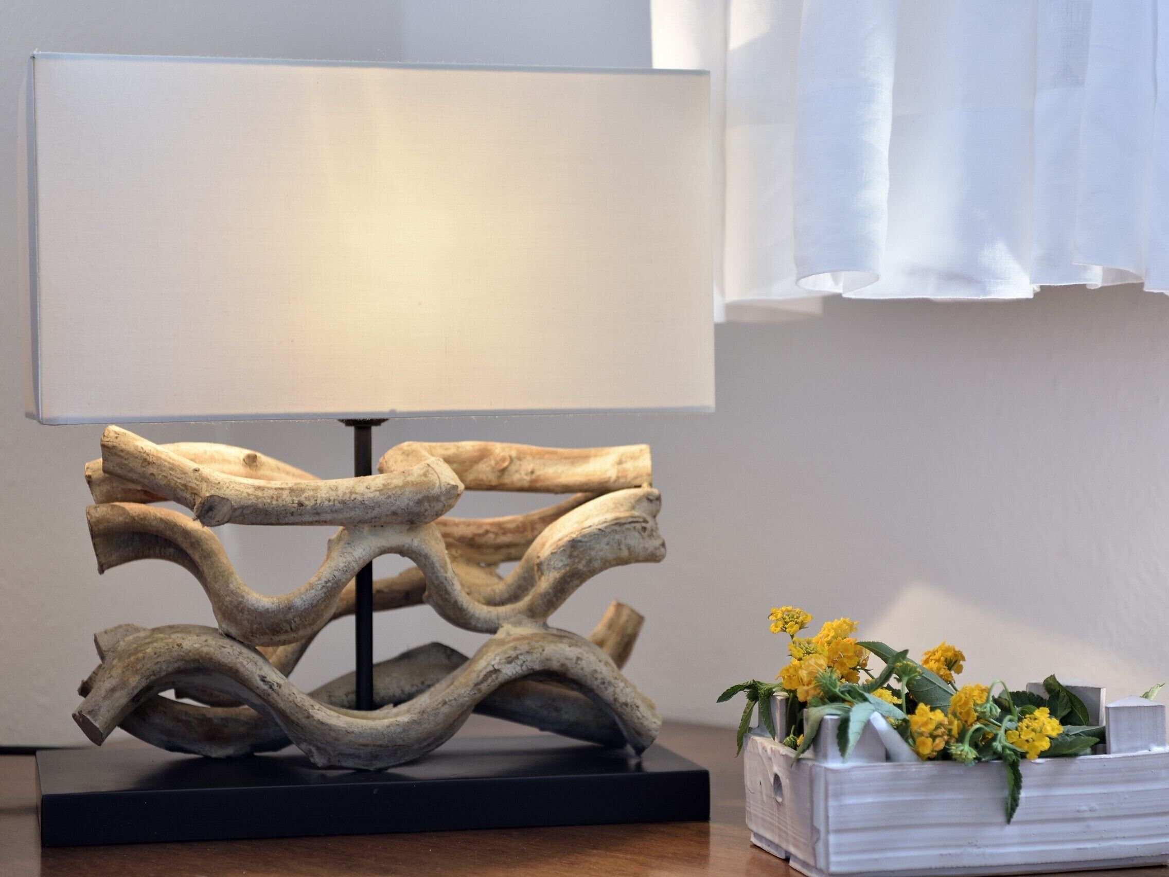 Eine moderne Lampe mit einem einzigartigen Holzdesign auf einem schwarzen Sockel, eine weiße Vase mit gelben Blumen und ein weißer Vorhang im Hintergrund.