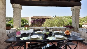 An outdoor dining area with stone pillars, a wooden pergola, and a view of a hillside with vegetation and buildings. - Holiday apartments L'Ea di Lavru 
SEA VIEW 3-rooms apartment-TOP INTERIOR DESIGN (Porto San Paolo)