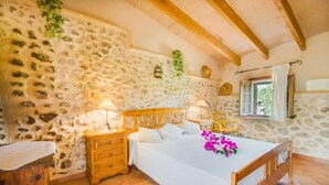 A bedroom with a stone wall, wooden bed frame, bedside tables, and a window with curtains. - Large rustic finca Can Nyany Buger in Mallorca (Buger)