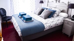 A neatly made bed with blue and white bedding, a chair, a bedside table with a lamp, and a patterned curtain. - Charming town house Avignon intra muros (Avignon)