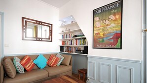 A living room with a beige sofa, colorful pillows, a wooden coffee table, a bookshelf, and a poster of Paris and Tokyo.