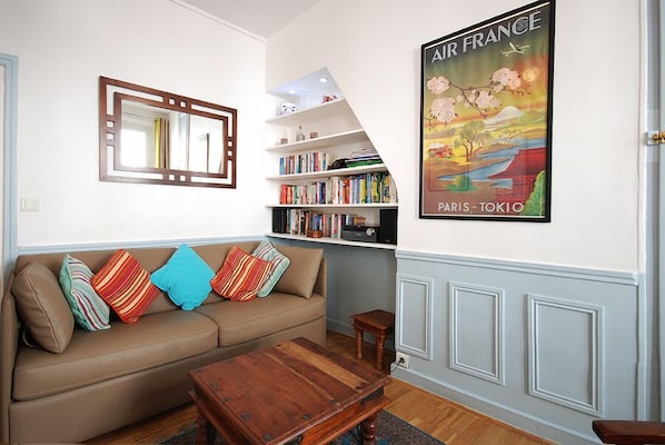 A living room with a beige sofa, colorful pillows, a wooden coffee table, a bookshelf, and a poster of Paris and Tokyo.