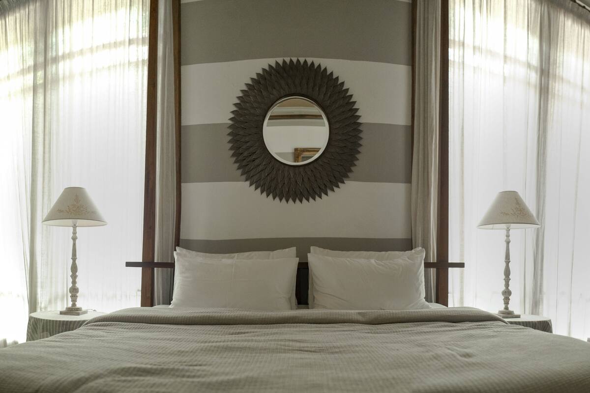 Habitación de hotel con una cama grande, dos mesitas de noche con lámparas, un espejo con marco de sol y cortinas traslúcidas.