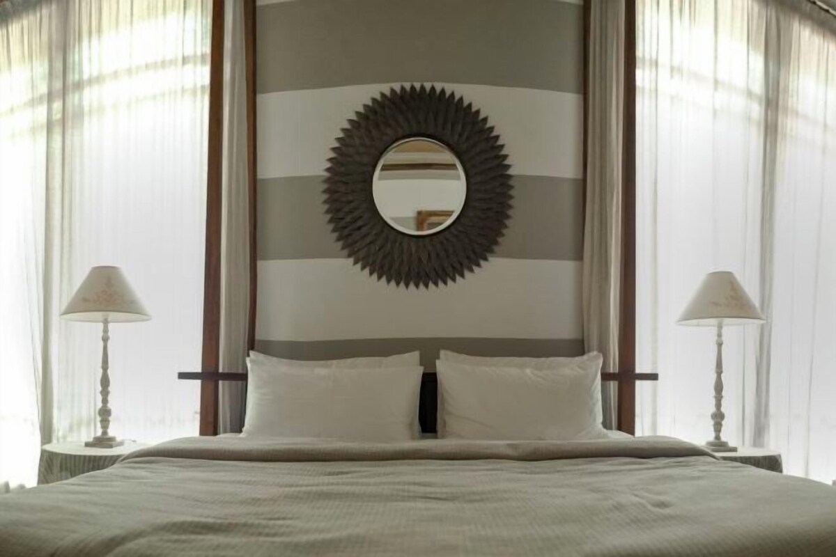 A hotel room with a bed, two bedside lamps, a large mirror with a sunburst frame, and a striped wall.