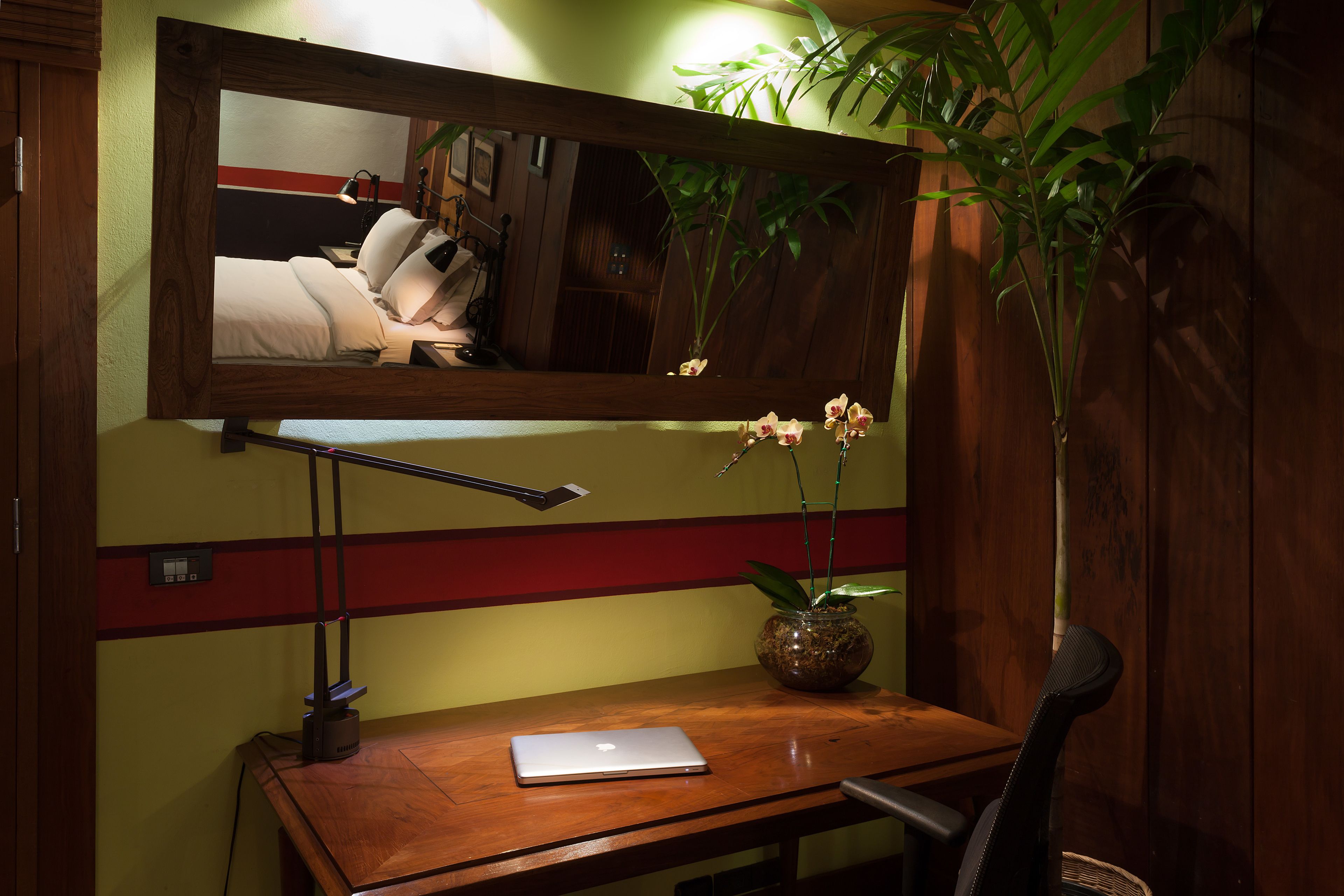 Un escritorio de madera con una computadora portátil, una lámpara de escritorio, un florero con una planta y un espejo que refleja una cama y una pared con rayas rojas y amarillas.