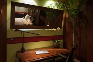 Un escritorio de madera con una computadora portátil, una lámpara de escritorio, un florero con una planta y un espejo que refleja una cama y una pared con rayas rojas y amarillas.