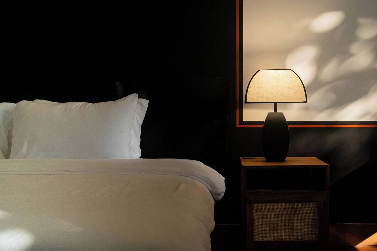 Una cama bien hecha con sábanas blancas, una mesita de noche con lámpara y un cuadro colgado en la pared.