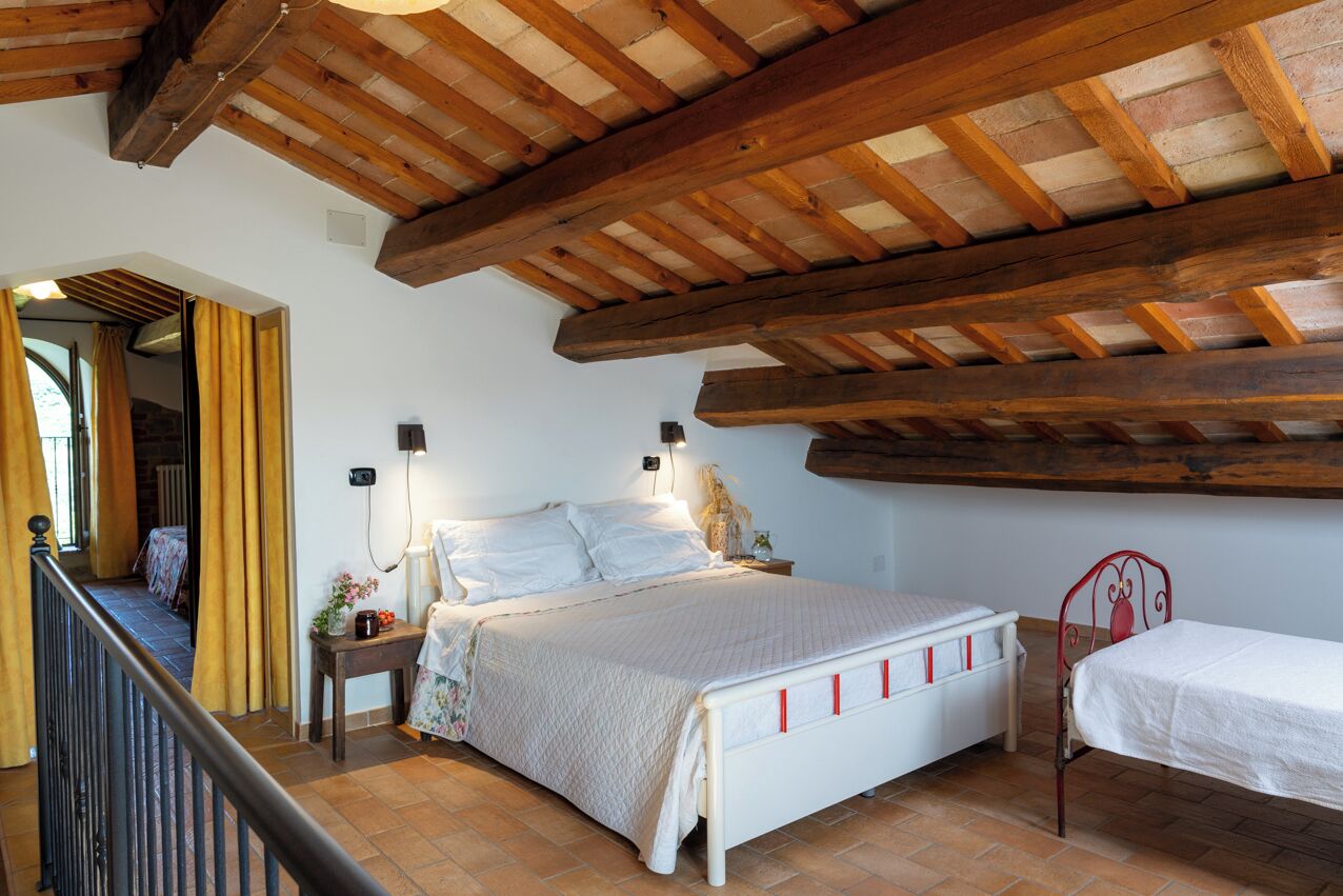 A bedroom with a wooden ceiling, a bed with a white headboard, a bedside table with a lamp, and a window with curtains.
