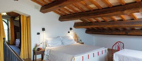 A bedroom with a wooden ceiling, a bed with a white headboard, a bedside table with a lamp, and a window with curtains.