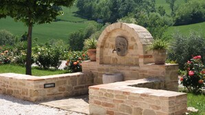 A stone-built outdoor water feature with a stone basin and a water spout, surrounded by a gravel area and potted plants.