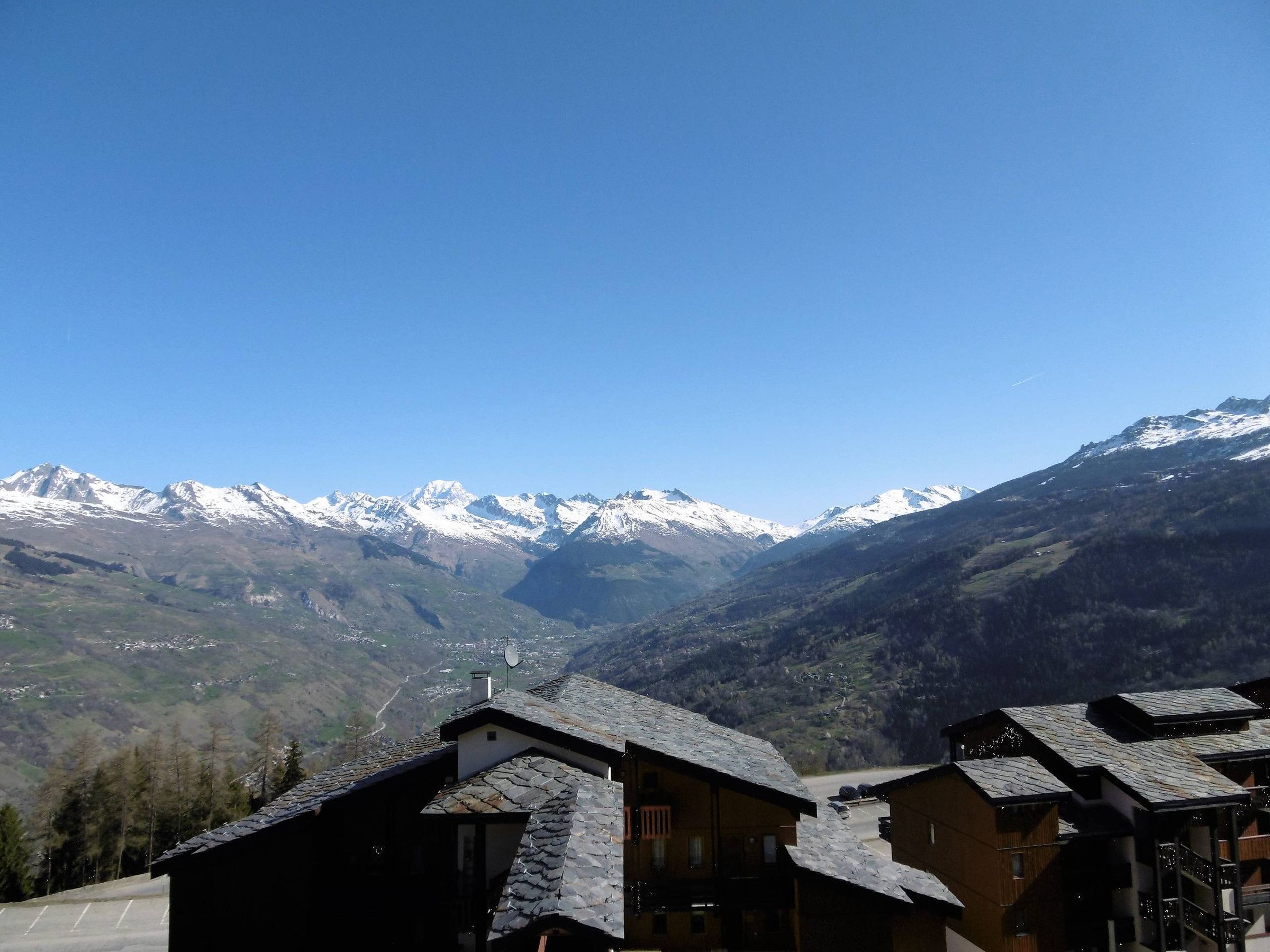 A mountain village with traditional wooden houses, a clear sky, and snow-capped peaks in the background.