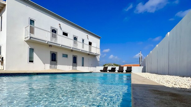 A swimming pool adjacent to a white building with a balcony, clear blue sky, and a sandy area. - Located only minutes away from the Palm Beaches. 2 bedroom 3 beds 2 bathroom (Noord)