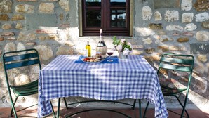 Una mesa con mantel de cuadros, una botella, un florero con flores y dos sillas frente a una pared de piedra con una ventana.