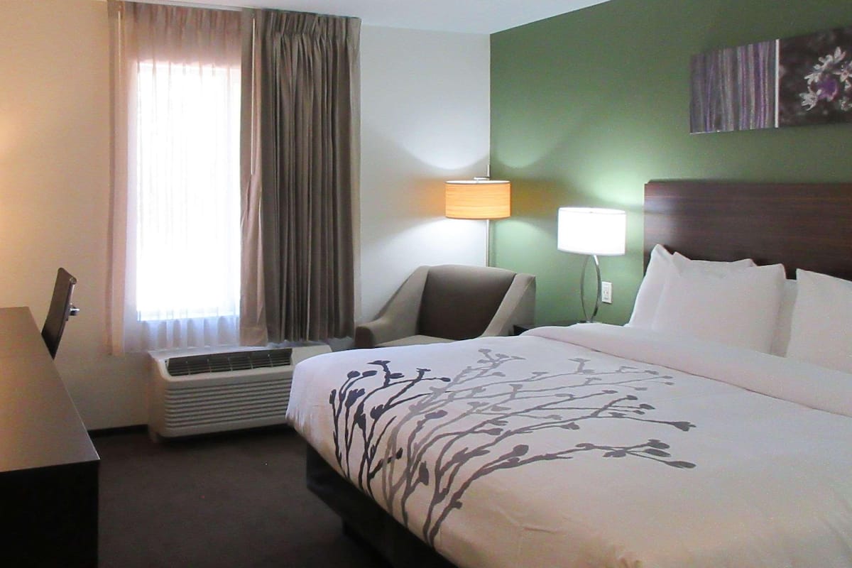 A hotel room with a bed, a chair, a desk, a lamp, and a window with curtains.
