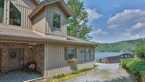 A two-story house with a stone entrance, a bench outside, and a view of a lake.