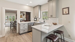 A modern kitchen with white cabinetry, a large island with bar stools, and a view of the backyard through an open door.