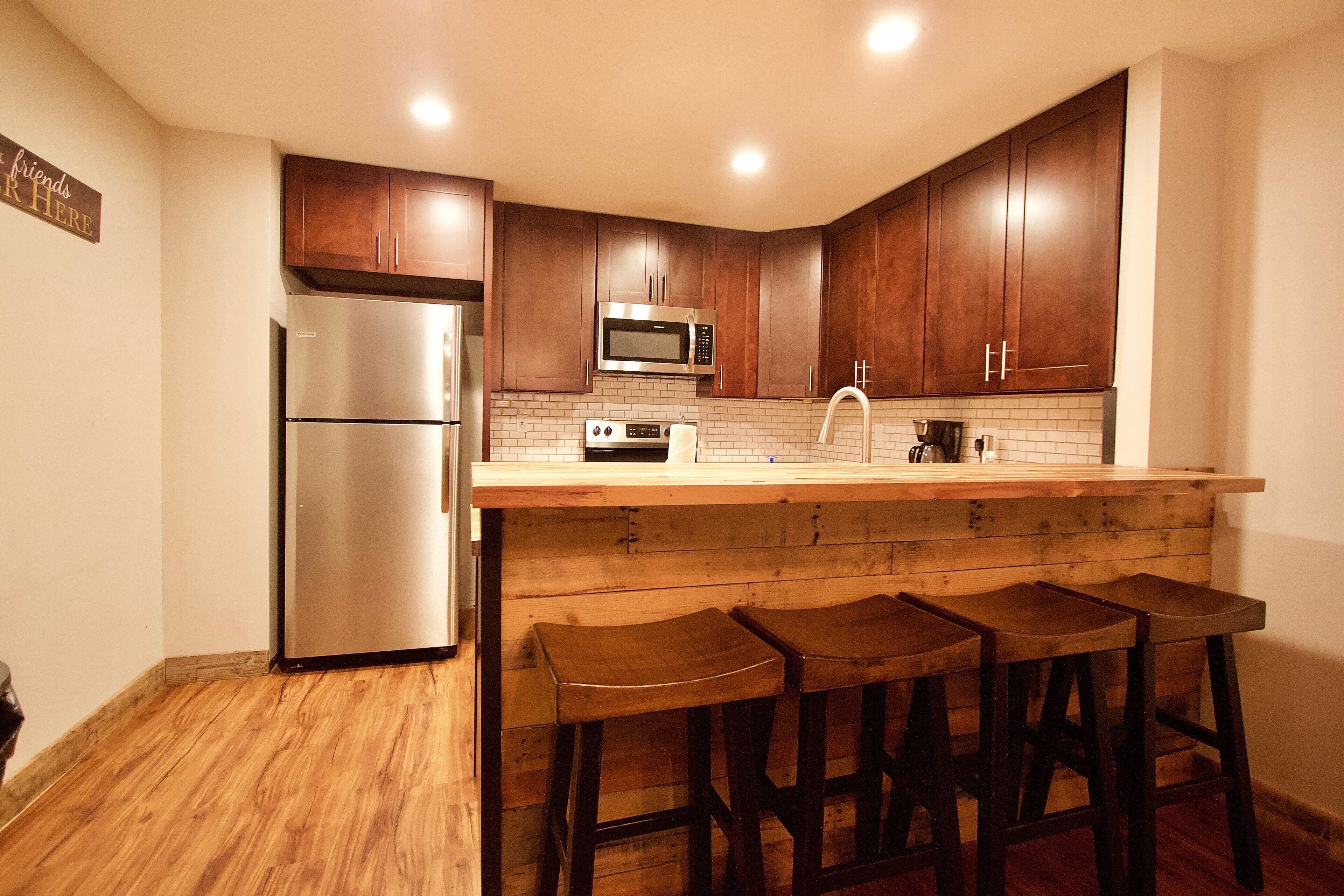 A kitchen with wooden cabinets, a stainless steel refrigerator, a microwave, and a wooden bar counter with stools.