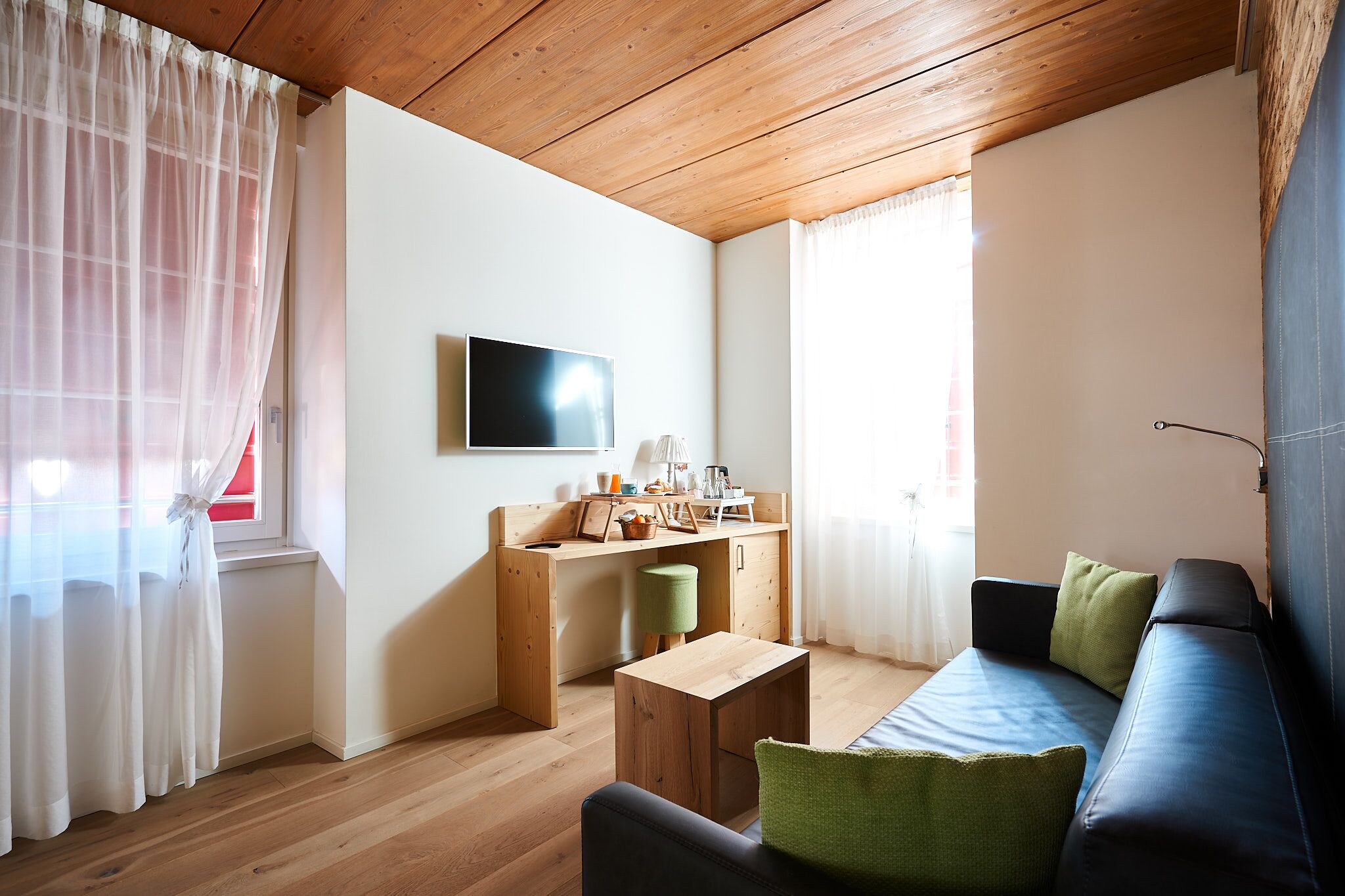 A compact living space with a wooden ceiling, a sofa with cushions, a small wooden table, a desk with a chair, and a TV mounted on the wall.