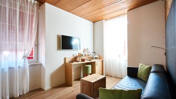 A compact living space with a wooden ceiling, a sofa with cushions, a small wooden table, a desk with a chair, and a TV mounted on the wall.