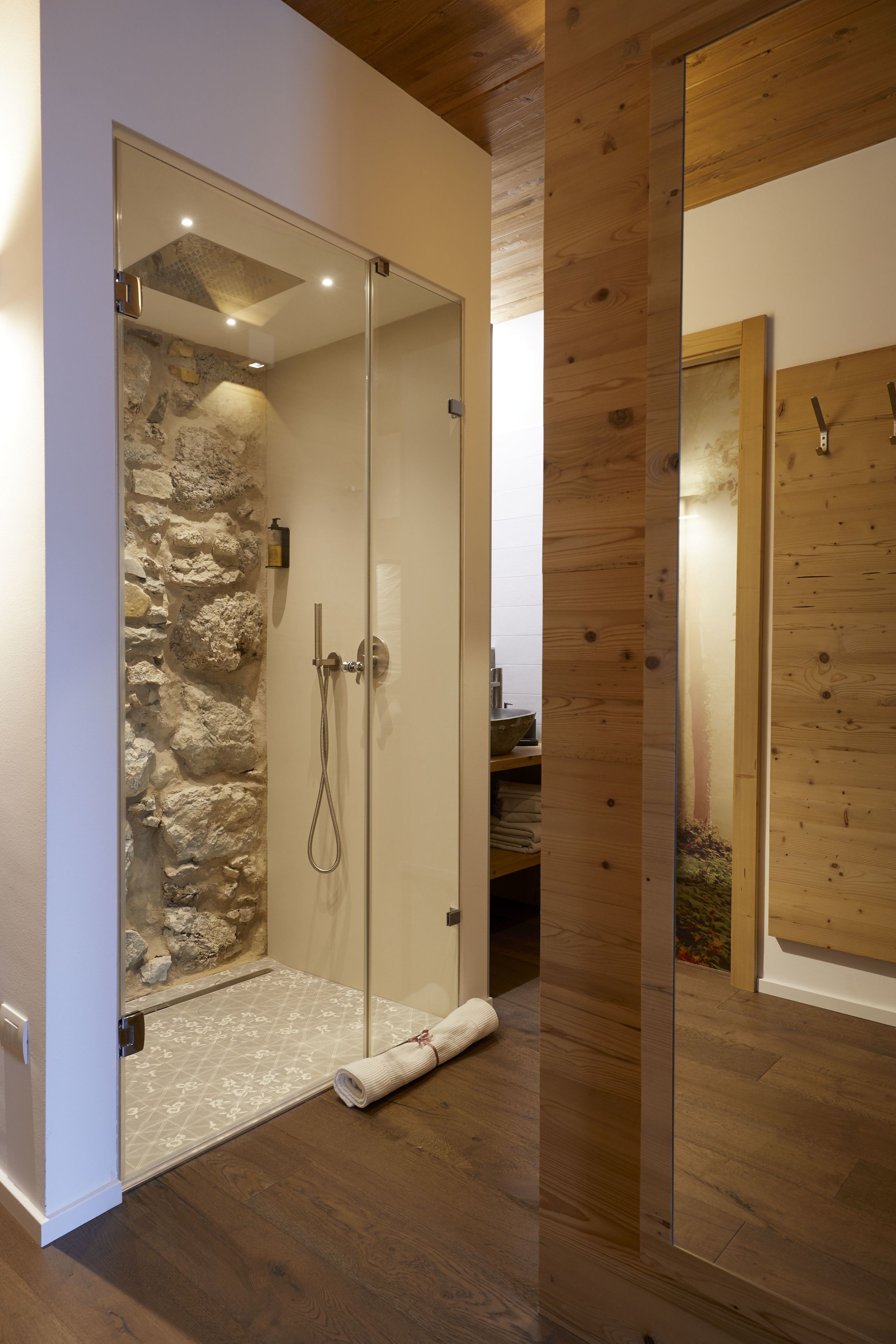A glass-enclosed shower with a stone wall, wooden flooring, and a towel on the floor.